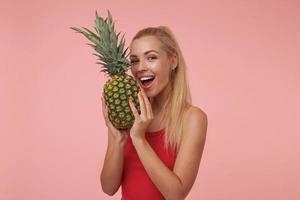 prise de vue en studio d'une jeune femme blonde avec une coiffure décontractée tenant de l'ananas dans ses mains, allant mordre l'ananas, regardant la caméra et souriant joyeusement photo