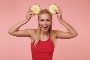 prise de vue en studio d'une charmante jeune femme aux longs cheveux blonds posant sur fond rose en chemise rouge, se moquant d'anneaux d'ananas frais, souriant joyeusement à la caméra photo