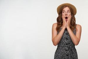 photo de studio d'une belle femme aux cheveux rouges étonnée en chapeau canotier regardant de côté avec de grands yeux et la bouche ouverte, tenant des paumes sur son visage et levant les sourcils, isolée sur fond blanc