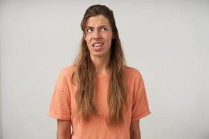 photo de studio d'une jeune femme aux cheveux longs debout sur fond blanc avec une coiffure décontractée et un maquillage naturel, regardant vers le haut et faisant la moue