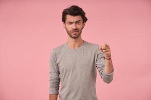 jeune homme barbu déconcerté avec une coupe de cheveux à la mode regardant la caméra et fronçant les sourcils, debout sur fond rose en pull gris, pointant vers l'avant avec index figer photo