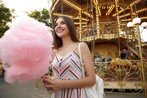 photo en plein air d'une jeune femme séduisante brune aux cheveux longs vêtue d'une robe d'été, posant sur un carrousel par temps chaud, tenant de la barbe à papa sur un bâton, regardant de côté avec un sourire sincère