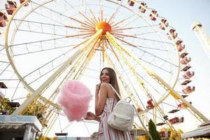 photo en plein air d'une jeune femme brune heureuse aux cheveux longs portant une robe romantique et un sac à dos blanc, debout au-dessus de la grande roue par une chaude journée d'été, tenant de la barbe à papa et souriant largement