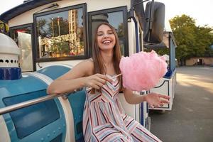 heureuse jeune femme charmante aux longs cheveux bruns assise près d'une voiture de train à vapeur dans un parc d'attractions, vêtue d'une robe d'été à bretelles et tenant un bâton avec de la barbe à papa rose, riant et regardant la caméra photo