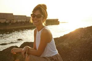 belle jeune femme aux cheveux noirs en bandeau et lunettes de soleil rencontrant l'aube sur le bord de mer, s'appuyant sur la pierre avec les mains croisées et souriant joyeusement à la caméra photo