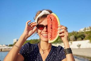 heureuse jeune femme brune en bandeau et lunettes de soleil vintage regardant joyeusement la caméra et tenant une tranche de pastèque par son visage, debout sur la vue sur la mer photo