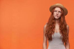 photo de studio d'une jolie jeune femme rousse en vêtements décontractés debout sur fond orange, portant une chemise grise et un chapeau de canotier, regardant pensivement de côté et mordant la lèvre inférieure