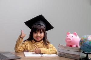 petite fille asiatique portant un bonnet de graduation avec une tirelire rose, économiser de l'argent, investir dans l'avenir, photo