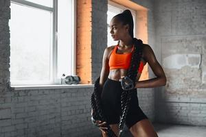 belle jeune femme africaine portant une corde de combat sur les épaules en se tenant debout dans la salle de sport photo