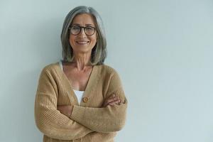 élégante femme senior gardant les bras croisés et souriant tout en se tenant contre le mur blanc photo