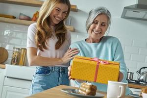 heureuse femme âgée recevant une boîte-cadeau de sa fille tout en passant du temps à la cuisine photo