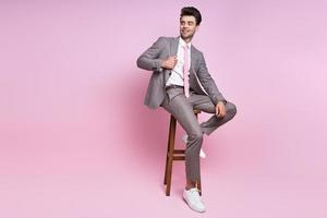 beau jeune homme en costume complet ajustant sa veste assis sur une chaise sur fond rose photo