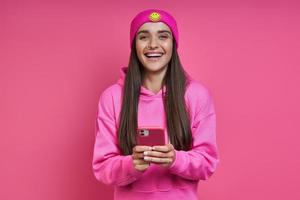 belle jeune femme en chemise à capuche et chapeau funky tenant un téléphone intelligent sur fond rose photo