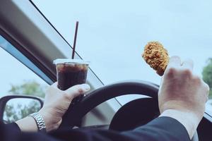 homme d'affaires conduisant une voiture en mangeant dangereusement du poulet frit et des boissons gazeuses froides photo
