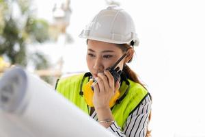 un ingénieur asiatique ou une jeune femme architecte met un casque pour la sécurité et regarde le plan pour inspecter le chantier de construction de l'usine de construction et utilise un talkie-walkie pour une conversation avec un entrepreneur.