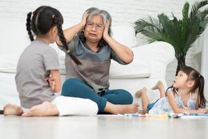 une vieille grand-mère âgée épuisée est assise par terre dans le salon et se sent mal fatiguée par les petits enfants qui courent et jouent fort, souffre de maux de tête, la nounou se sent épuisée par les enfants bruyants photo