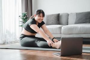 femme potelée asiatique assise sur le sol dans le salon pratique une leçon de yoga en ligne avec l'ordinateur. femme ayant un cours de formation à la méditation sur l'ordinateur portable. photo
