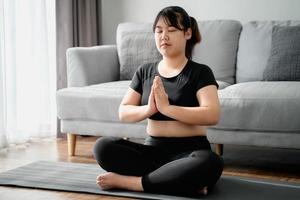 femme potelée asiatique assise sur le sol dans le salon pratique le cours de yoga. femme ayant un cours de formation à la méditation. photo
