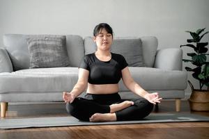 femme potelée asiatique assise sur le sol dans le salon pratique le cours de yoga. femme ayant un cours de formation à la méditation. photo