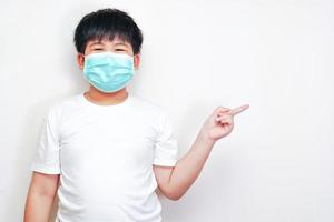 le garçon asiatique portant un masque chirurgical médical, portant un t-shirt blanc et pointant son doigt sur le côté. photo