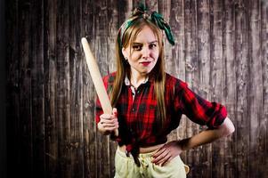 jeune femme au foyer drôle en chemise à carreaux et short jaune style pin up avec rouleau à pâtisserie de cuisine sur fond en bois. photo