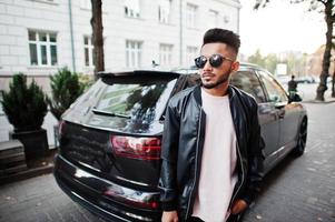 homme de barbe indien élégant à la veste en cuir noir et lunettes de soleil contre la voiture suv d'affaires. modèle indien posé en plein air dans les rues de la ville. photo