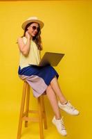 femme élégante positive en chapeau et robe d'été travaillant avec un ordinateur portable sur fond jaune photo