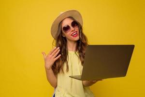 fille branchée élégante avec des cheveux ondulés dans des lunettes de soleil et un chapeau appelle un appel vidéo avec un ordinateur portable avec des émotions heureuses photo