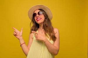 femme brune heureuse en robe jaune et chapeau lève les yeux, pointant du doigt et sourit sincèrement sur fond jaune. photo