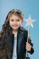 une petite fille satisfaite a de longs cheveux bouclés, vêtue d'une veste en cuir noir, porte une couronne, tient une baguette magique, isolée sur fond bleu. un enfant positif aux cheveux noirs anticipe un miracle photo