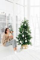 photo verticale d'une femme positive pose avec un chien jack russell terrier, vêtue de vêtements domestiques décontractés, pose ensemble dans la chambre avec un arbre de nouvel an décoré, anticipez les vacances. heure d'hiver