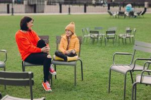 photo d'une jeune mère et d'une fille sympathiques et positives qui posent à l'extérieur dans une zone de loisirs avec une pelouse verte sur des chaises, vêtues d'une tenue à la mode, rigolent à quelque chose de drôle. famille et communication