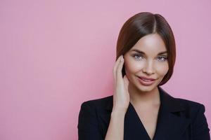 photo d'une charmante femme entrepreneure européenne à la peau saine, touche doucement le visage, revient du salon de spa, porte une tenue noire formelle, regarde avec confiance la caméra, isolée sur fond rose