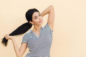 une femme pensive aux cheveux noirs fait une queue de cheval, a des lèvres pleines et une apparence agréable, porte une tenue décontractée, se tient sur fond de studio, concentrée à distance. concept de personnes, de beauté et de style de vie photo