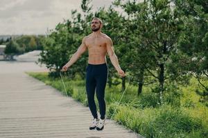 un homme sportif à la barbe épaisse fait des exercices avec une corde à sauter, a un torse musclé, reste en bonne forme physique, pose à l'extérieur. remise en forme et concept sain. coureur masculin se réchauffe avec un équipement de sport photo