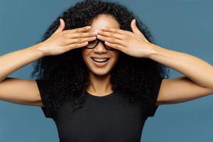 femme secrète aux cheveux bouclés, couvre les yeux, porte des lunettes, attend la surprise, vêtue d'un t-shirt décontracté noir, pose sur fond bleu, a une manucure anticipant le miracle. la femme se cache le visage photo