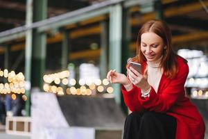 jeune femme européenne satisfaite en manteau rouge, tient un téléphone portable, lit la notification, connectée à Internet sans fil, a du temps libre, pose à l'extérieur, attend quelqu'un, aime la communication en ligne photo