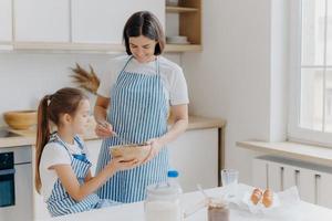 famille heureuse à la cuisine. une jolie femme et sa fille préparent la boulangerie ensemble, portent des tabliers, aiment cuisiner ensemble, profitent de l'atmosphère domestique, s'amusent à l'intérieur. enfants, maternité, concept de cuisson photo