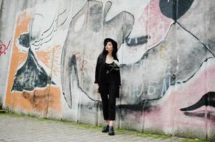 fille sensuelle tout en noir, lèvres rouges et chapeau. une femme dramatique gothique tient une fleur de chrysanthème blanche contre un mur de graffitis. photo