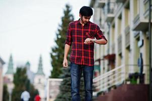 jeune étudiant indien en chemise à carreaux rouge et jeans posés à la ville et regardant ses montres. photo