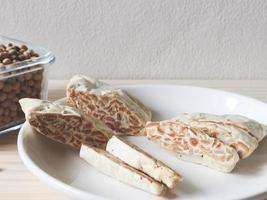 tempeh cru ou tempe. tranches de tempeh dans une assiette en céramique blanche et un bol de graines de soja sèches sur une table en bois. le tempeh est un aliment traditionnel indonésien. photo