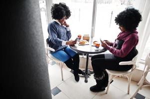 deux femmes afro-américaines aux cheveux bouclés portent des chandails assis au café intérieur avec des téléphones à portée de main. photo