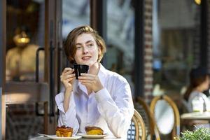 femme caucasienne sirotant un café expresso chaud tout en étant assise à l'extérieur du café-bistro de style européen profitant de la vie lente avec l'ambiance matinale sur la place de la ville avec une pâtisserie sucrée