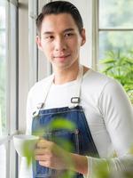 jardinier adulte jeune homme asiatique une personne beau debout pense regarder et souriant se détendre heureux de boire une tasse de café dans le café de la chambre avec fenêtre en verre et arbres verts au printemps matin. photo