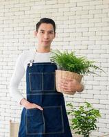 portrait jardinier jeune homme asiatique beau une personne debout souriant regardant la main tenant le panier pot petit arbre feuille verte dans un atelier calme maison plante mur blanc. passe-temps travail heureux et concept de soins photo