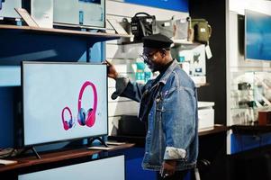 homme afro-américain décontracté élégant à la veste en jean et au béret noir près du nouvel écran de télévision au magasin d'électronique. photo