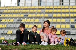 jeune mère élégante avec quatre enfants assis sur l'herbe contre le stade. la famille sportive passe du temps libre à l'extérieur avec des scooters et des patins. photo