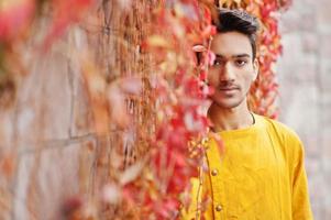 homme élégant indien en vêtements traditionnels jaunes posés en plein air contre le mur de feuilles rouges. photo