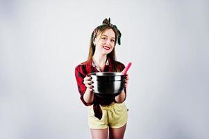 jeune femme au foyer drôle en chemise à carreaux et short jaune pin up style avec casserole et cuillère de cuisine isolé sur fond blanc. photo