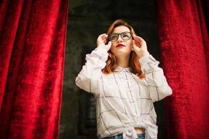 jolie femme rousse à lunettes, porter sur un chemisier blanc posant à l'arche de rideaux rouges ouverts. photo